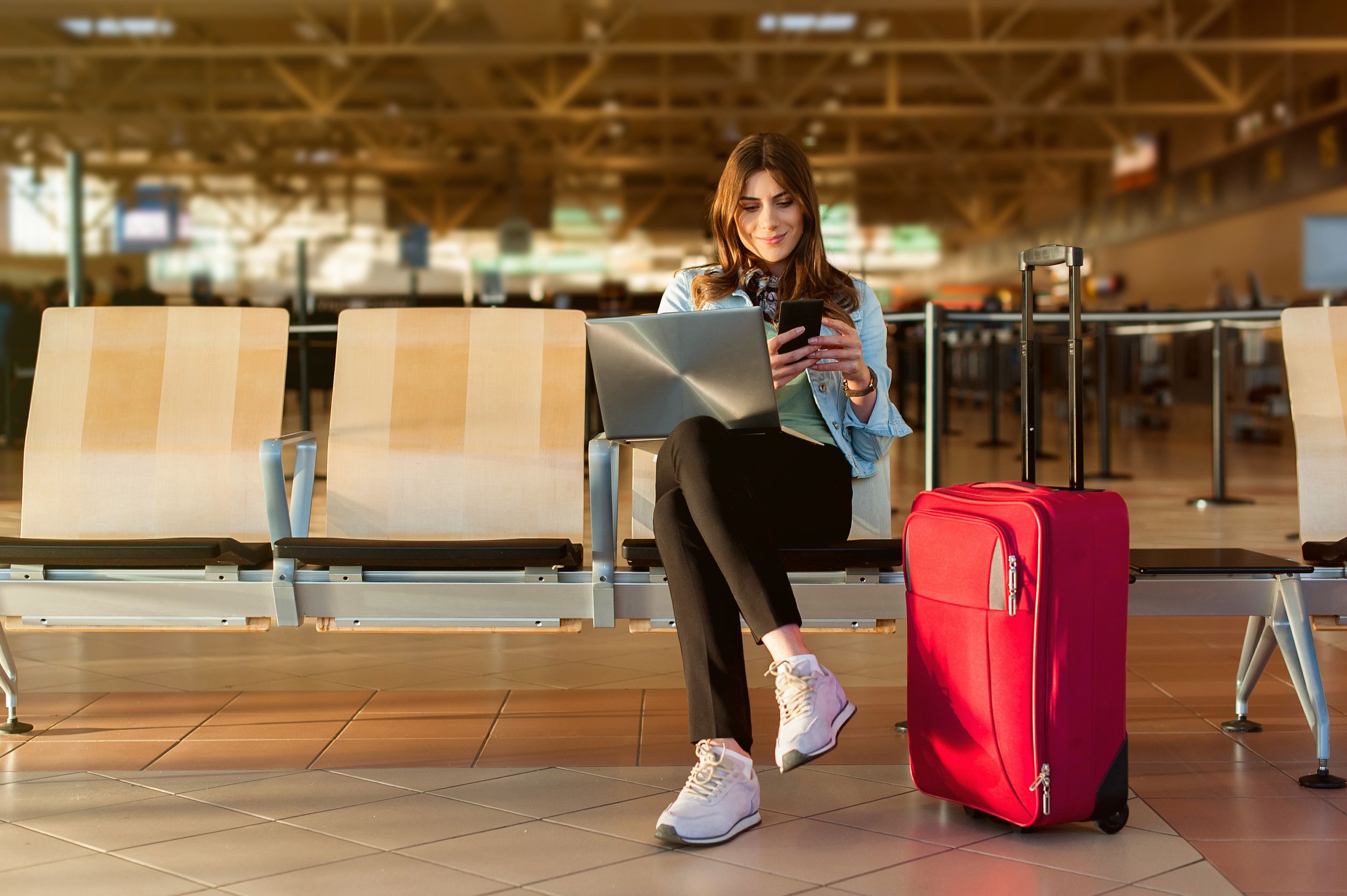 Giovane passeggera dell'aeroporto con smartphone e computer portatile seduta nella sala del terminal in attesa Giovane passeggera dell'aeroporto con smartphone e computer portatile seduta nella sala del terminal in attesa