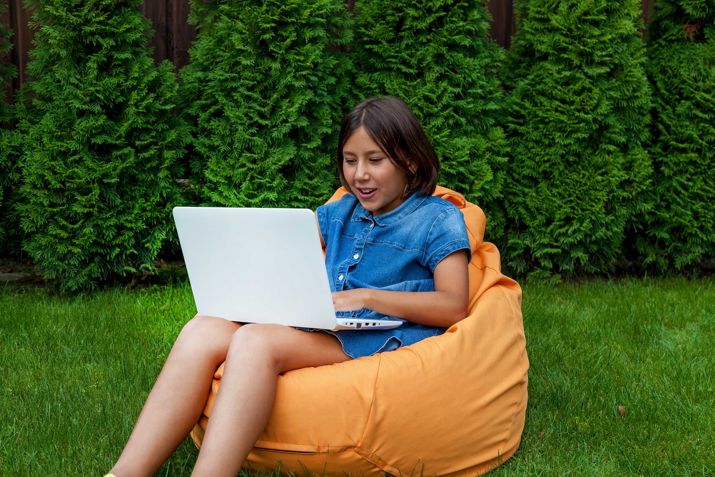 Bambina seduta su un cuscino con un computer portatile aperto sull'erba verde nel cortile della casa Bambina seduta su un cuscino con un computer portatile aperto sull'erba verde nel cortile della casa